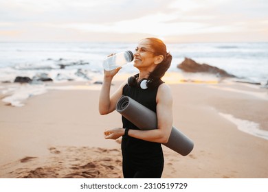 Smiling mature european woman in sportswear with mat, drinks bottle of water, enjoy workout in morning on sea beach, outdoor. Weight loss sport, fitness at sunrise and body care - Shutterstock ID 2310519729