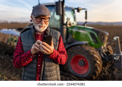 Smiling mature farmer looking at mobile phone in front of tractor in field. Smart farming concept - Shutterstock ID 2244074381
