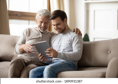 Smiling mature father and grownup son relax on sofa in living room have fun using modern tablet gadget together. Happy elderly dad and adult man child watch video browse internet on pad at home. - Powered by Shutterstock