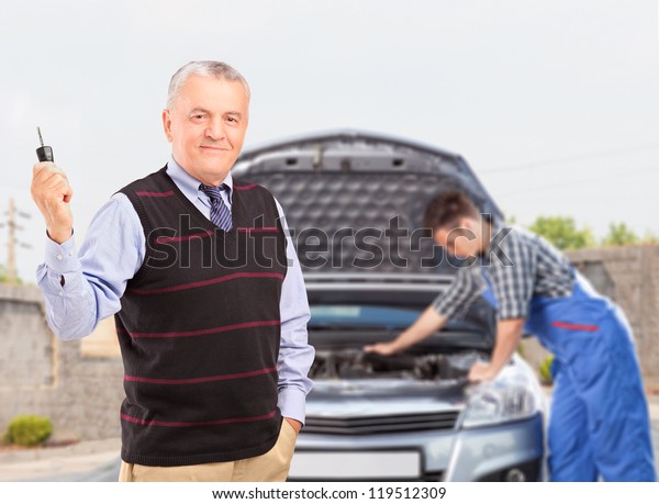 Smiling mature gentleman holding a\
car key while in the background mechanic is checking his\
car
