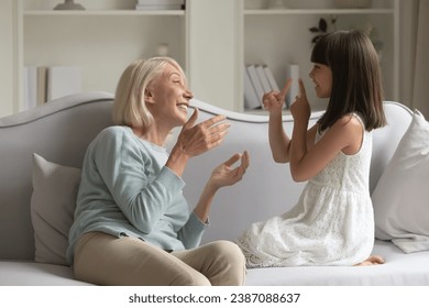 Smiling mature grandmother and little granddaughter practicing sign language, sitting on cozy sofa at home together, happy senior granny and adorable grandchild girl speaking, communicating - Powered by Shutterstock