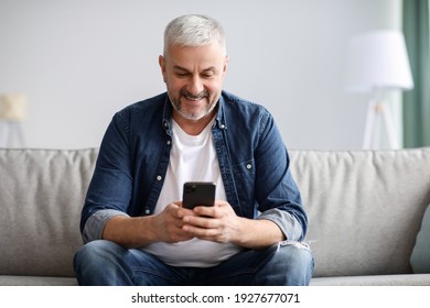 Smiling Mature Grey-haired Man Sitting On Couch And Using Mobile Phone At Home, Copy Space. Cheerful Middle-aged Man With Smartphone, Chatting With His Family Or Using Mobile Application