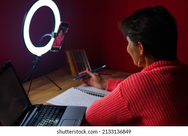 Smiling Mature Hispanic Woman Making A Live Stream Video With A Ring Light, Her Phone And Laptop. Creative Blogging Set Up For Drawing And Coloring Art Tutorial.