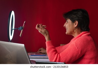 Smiling Mature Hispanic Woman Making A Live Stream Video With A Ring Light, Her Phone And Laptop. Creative Blogging Set Up For Drawing And Coloring Art Tutorial.