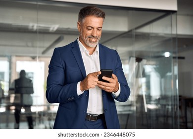 Smiling mature Latin or Indian businessman holding smartphone in office. Middle aged manager using cell phone mobile app. Digital technology application and solutions for business success development.