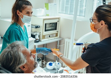 Smiling Mature Male Patient Sitting On Dentist's Chair While Female Dentist And Dental Assistant In Protective Face Mask And Gloves Taking Teeth Impression In Dental Clinic
