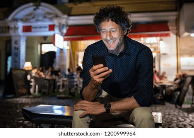 Smiling Mature Man Checking Phone While Sitting On Bench Outside Restaurant. Successful Businessman Reading Message On Mobile Phone At Night. Senior Entrepreneur Feeling Happy On Receiving Mail.