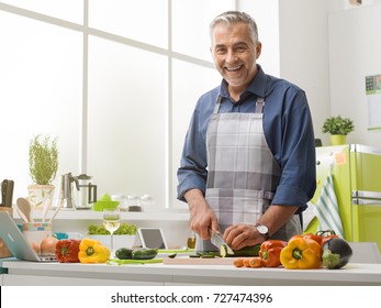 Smiling Mature Man Cooking In The Kitchen At Home, He Is Wearing An Apron And Slicing Fresh Vegetables, Healthy Lifestyle Concept