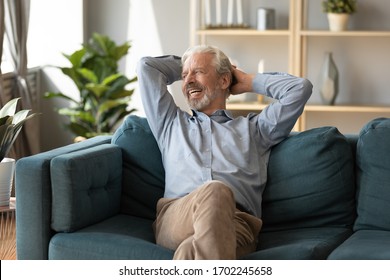 Smiling mature man dreaming, relaxing on cozy couch alone, happy older senior male with hands behind head sitting resting on sofa in living room, looking in window, thinking about good future