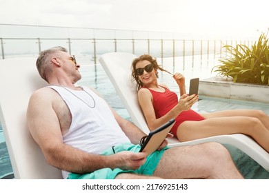 Smiling Mature Man Flirting With Woman In Red Swimsuit Sitting On Nearby Chaise-lounge