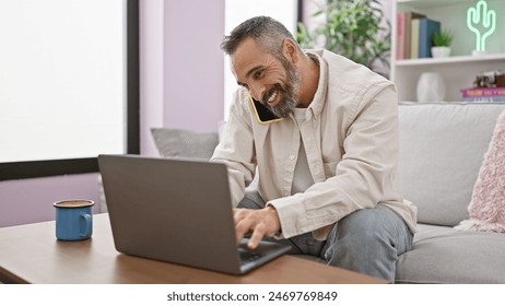 Smiling mature man with grey beard multitasking with phone and laptop in a cozy living room. - Powered by Shutterstock