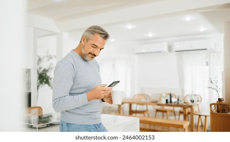 Smiling mature man with grey hair enjoying a cup of coffee and smartphone in a cozy kitchen setting - Powered by Shutterstock