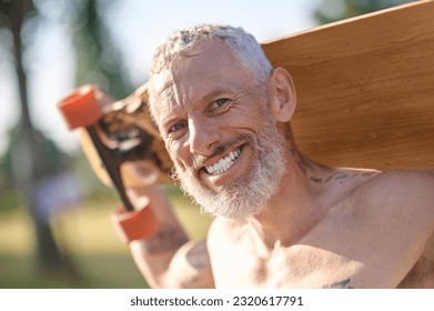Smiling mature man holding longboard in a city park.