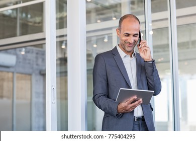 Smiling Mature Man Looking At Digital Tablet While Talking Over Phone. Mature Businessman In A Happy Conversation Over Cellphone. Successful Senior Business Man In Formal Talking Over Phone.