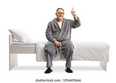 Smiling Mature Man In Pajamas And A Robe Sitting On A Bed And Pointing Up Isolated On White Background