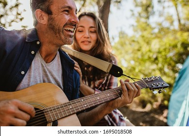 Smiling Mature Man Playing Guitar With A Woman Sitting By. Happy Mature Couple Camping In Nature.