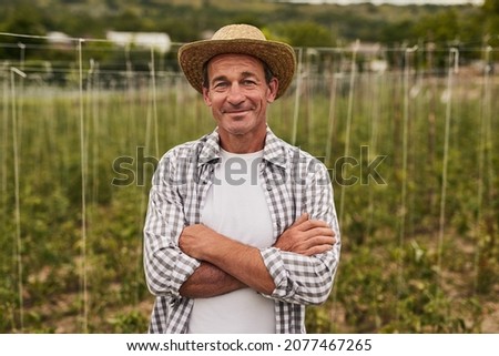 Smiling mature man in straw hat and plaid shirt folding arms while standing on own farm and looking at camera