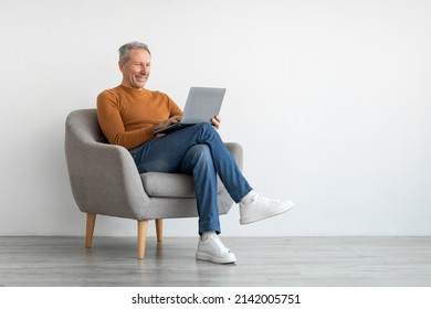 Smiling mature man using laptop sitting on armchair in living room. Happy confident senior male adult resting at home working on pc, typing on keyboard isolated on white studio wall, full body length