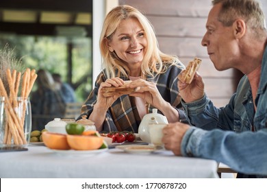 Smiling mature man and woman are having toasts with vegetables and hot drinks on terrace in countryside