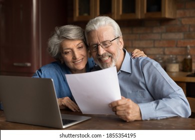 Smiling Mature Married Couple Reading Letter With Good News, Using Laptop, Sitting At Table In Kitchen, Happy Grey Haired Wife Hugging Senior Husband Holding Paper Sheet, Document, Money Refund