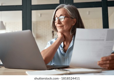 Smiling mature middle aged business woman using laptop working on computer sitting at desk. Happy old businesswoman hr holding cv interviewing distance applicant, senior seeker searching job online.