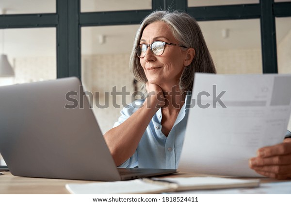 Smiling mature middle aged business woman using\
laptop working on computer sitting at desk. Happy old businesswoman\
hr holding cv interviewing distance applicant, senior seeker\
searching job online.