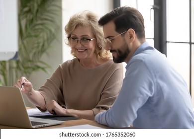 Smiling mature middle aged businesswoman mentor coach teaching intern, pointing at laptop screen, helping with corporate software to new employee, giving instructions, colleagues working together