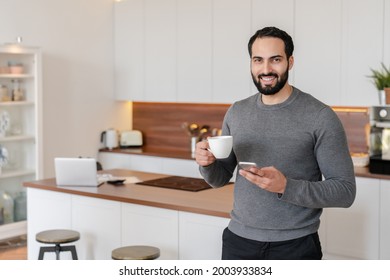 Smiling mature Middle Eastern arabian man using smart phone during breakfast drinking hot beverage at home in the kitchen. Good connection, checking social media, loading photos on profile, blogger - Powered by Shutterstock