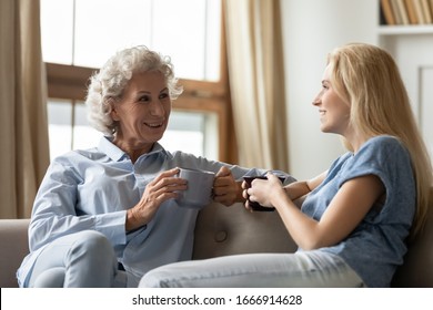 Smiling Mature Mother And Adult Daughter Sit Rest On Sofa In Living Room Chatting Talking Drinking Tea Or Coffee, Happy Senior Mom And Grownup Girl Child Relax On Couch At Home, Enjoy Time Together