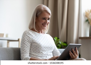 Smiling mature old woman holding using digital tablet relaxing sit on sofa reading e book in reader app, happy middle aged senior grandma browsing internet shopping online on pad computer at home - Powered by Shutterstock