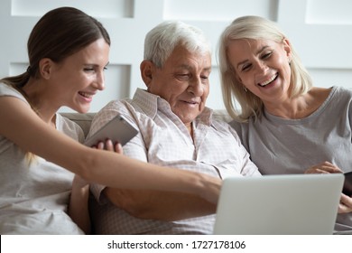 Smiling Mature Parents Sit Relax On Couch With Grownup Daughter Using Modern Laptop Gadget Together, Happy Senior Mom And Dad Rest Have Fun Watch Video With Adult Millennial Girl On Computer