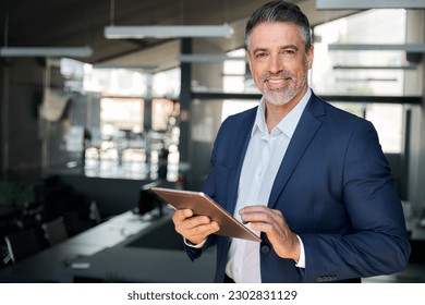 Smiling mature professional business man, happy middle aged successful company ceo, confident executive wearing blue suit standing in office holding tablet computer corporate technology. Portrait - Powered by Shutterstock