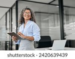 Smiling mature professional business woman bank manager, older happy female executive or lady entrepreneur holding digital tablet pad standing in office at work, looking away at copy space.