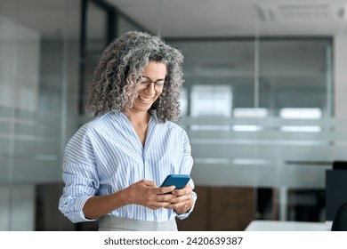 Smiling mature professional business woman entrepreneur using smartphone in office. Happy older senior female executive, middle aged businesswoman looking at mobile cell phone standing at work. - Powered by Shutterstock