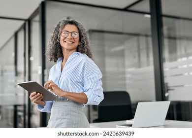 Smiling mature professional business woman bank manager, older happy female executive or lady entrepreneur holding digital tablet pad standing in office at work, looking away at copy space. - Powered by Shutterstock
