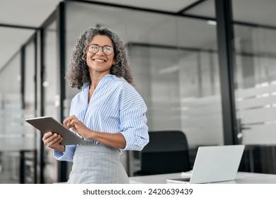 Smiling mature professional business woman bank manager, older happy female executive or lady entrepreneur holding digital tablet pad standing in office at work, looking away at copy space.