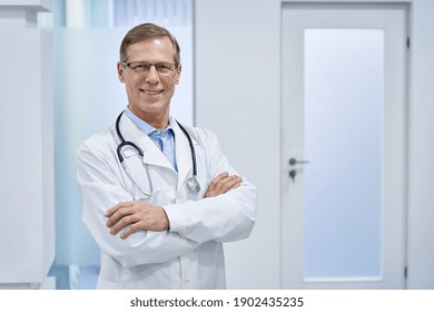 Smiling Mature Senior Doctor Wearing Glasses, Stethoscope And White Lab Medical Coat Standing In Hospital Looking At Camera. Middle Aged Old 60s Male Physician, Practitioner, Cardiologist Portrait.