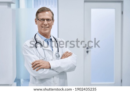Smiling mature senior doctor wearing glasses, stethoscope and white lab medical coat standing in hospital looking at camera. Middle aged old 60s male physician, practitioner, cardiologist portrait.