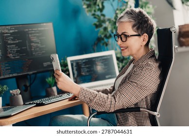 A smiling mature software developer works in a bright home office, reviewing code on laptop and monitors while using her smartphone for communication, conveying remote work, productivity and modern technology.