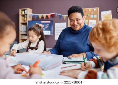 Smiling mature teacher of nursery school looking at one of learners drawing with crayons while sitting among intercultural kids - Shutterstock ID 2228718051
