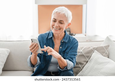 Smiling mature woman in casual clothes scrolling social media while relaxing on the couch at home. Middle-aged freelancer using cellphone for e-learning e-business - Powered by Shutterstock