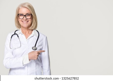 Smiling Mature Woman Doctor With Stethoscope Pointing At Copy Space Advertise Medical Health Care Services Pharmacy Clinic, Happy Old Lady Professional Nurse Isolated On White Grey Studio Background