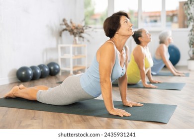 Smiling mature woman doing cobra pose of yoga on black mat in light training room - Powered by Shutterstock