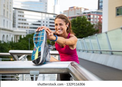 Smiling Mature Woman Doing Stretching With Her Leg Raised. Fitness Woman Listening To Music With Earphones While Stretching Leg In The City. Beautiful Runner Warming Up.