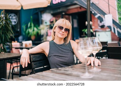 Smiling Mature Woman In Eyeglasses Sitting On Couch In Bar Outdoors With Wine Glasses And Blurry Restaurant Background, Drinking White Wine, Eating Cheese. Summer Sunny Day On Patio. People Lifestyle