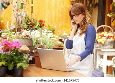 Smiling Mature Woman Florist Small Business Flower Shop Owner.  She Is Using Her Telephone And Laptop To Take Orders For Her Store. Shallow Focus.