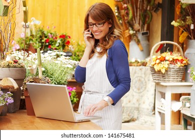 Smiling Mature Woman Florist Small Business Flower Shop Owner.  She Is Using Her Telephone And Laptop To Take Orders For Her Store. Shallow Focus.