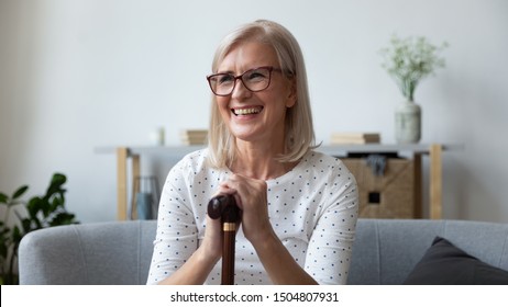 Smiling mature woman in glasses holding hands on wooden cane, happy disabled female sitting on couch at home, using walking stick during rehabilitation, older people healthcare concept - Shutterstock ID 1504807931