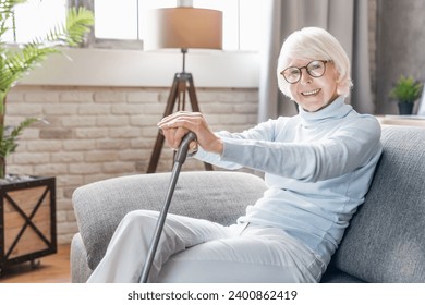 Smiling mature woman in glasses holding cane while sitting on sofa at home looking at camera. Seniors lifestyle. Lonely grandmother with walking disability, moving impairment - Powered by Shutterstock