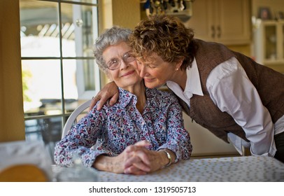 Smiling Mature Woman Greets Her Cheerful Elderly Mother As She Sits At The Kitchen Table.